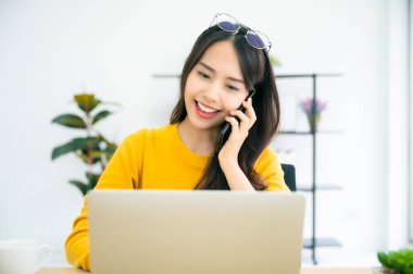 Evde çalışmak, video konferansı, çevrimiçi görüşme videosu bilgisayar ekranına bakan Asyalı genç bir kadın evde bilgisayar başında çalışıyor. Arkadaşlarıyla cep telefonu konuşmaları yapıyor.