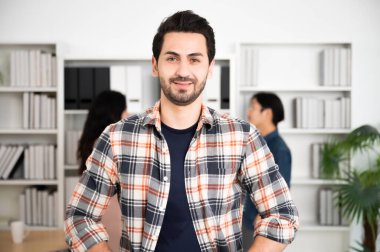 Genç Asyalı kendine güvenen takım lideri kameraya bakıyor ve arka plandaki bulanık insanlarla modern ofis odasında gülümsüyor.