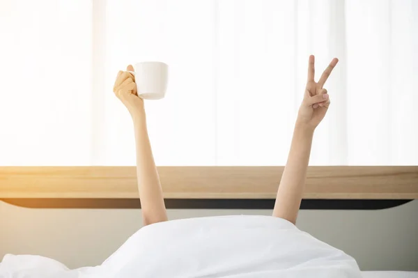 Woman waking up in the morning on bed. She hiding under the blanket and stretching out two arms with a cup of coffee and showing peace sign