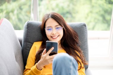 Mutlu genç Asyalı kadın evde dinleniyor. Kanepede oturuyor ve cep telefonu kullanıyor.