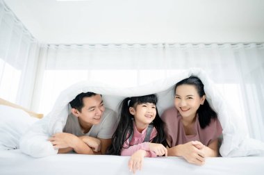 Mutlu Asyalı aile bireyleri yatak odasında birlikte vakit geçiriyorlar. Baba ve anne ve kızı yatakta dinleniyor ve eğleniyorlar.