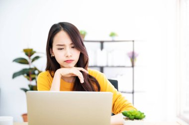 Asyalı genç bir kadın ciddi ciddi evdeki bilgisayar dizüstü bilgisayarında çalışıyor. Çözüm bulma sorununu düşünüyor.