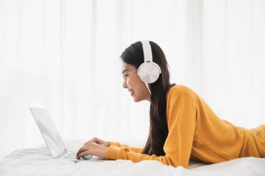 Güzel Asyalı genç kadın kulaklık takıyor ve bilgisayar kullanıyor. Yatak odasında boş vakit geçirir ve cihaz ekranını izlemekten zevk alır.