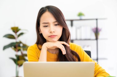 Asyalı genç bir kadın ciddi ciddi evdeki bilgisayar dizüstü bilgisayarında çalışıyor. Çözüm bulma sorununu düşünüyor.