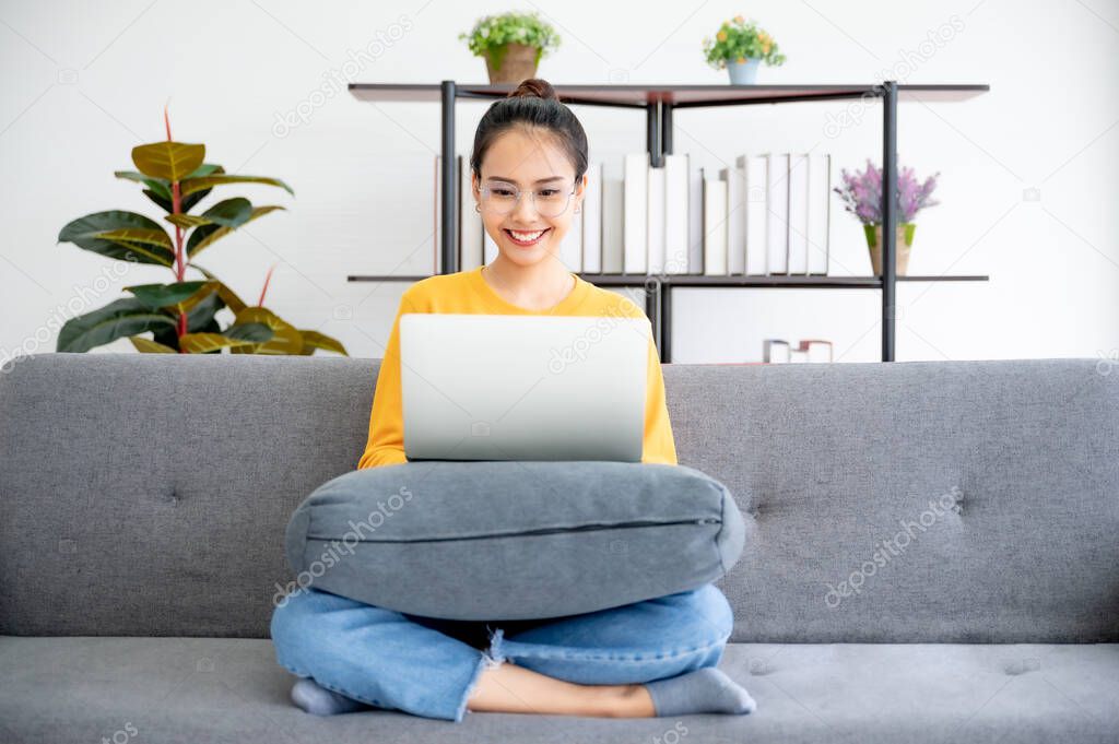 Asian woman wear glasses feeling happy and smile. She working on computer laptop and sitting on sofa at living room