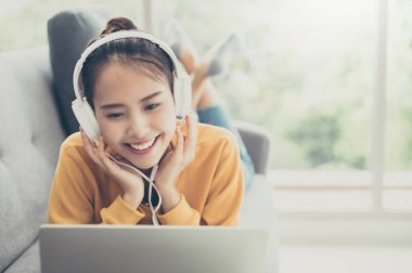 Laptop kullanan, kulaklık takan, müzik dinleyen ve oturma odasında kanepede uzanan güzel Asyalı bir kadın.