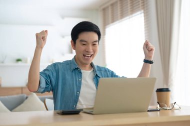 Asyalı bir adamın kazanmasını ya da bilgisayar laptopunda ecommerce alışveriş teklifi almasını kutlayan komik bir coşku. Heyecanlı mutlu çocuk kazanan başarıyı kutlayan deftere bakıyor.