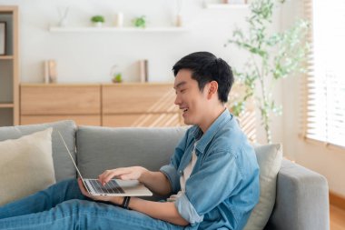Genç Asyalı adam rahat koltukta dinleniyor, evde dizüstü bilgisayar kullanıyor, sosyal ağda arkadaşlarıyla sohbet ediyor, haftasonunu geçiriyor, film izliyor, online alışveriş yapıyor.