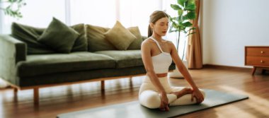 Genç Asyalı kadın evde nilüfer pozisyonunda meditasyon yapıyor, spor kıyafetleriyle yerde oturuyor. Rahatlama, huzur ve farkındalık için nefes alma egzersizi.
