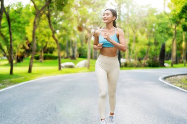 Asyalı bir kadın halka açık bir parkta koşuyor, atletik kıyafetler giyiyor ve yeşil, doğal bir ortamda kardiyo egzersizinin tadını çıkarıyor.