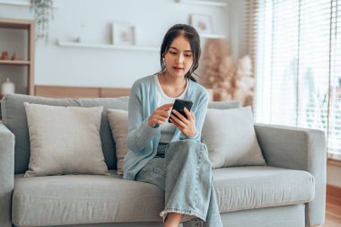 Oturma odasında komik mobil uygulamalar kullanarak rahat bir koltukta oturan gülümseyen genç Asyalı kadın. Evdeki Kadın, Çevrimiçi Alışveriş Yapıyor, Arkadaşlarına Mesajlaşıyor, Sosyal Medya 'ya Gönderiyor