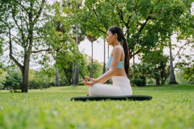 Barışçıl bir park ortamında meditasyon yapan Asyalı kadın, güneşli bir günde arka planda ağaçlarla yeşil çimlerde bacak bacak üstüne atarak oturuyor. Farkındalık, kişisel bakım ve açık hava rahatlığı kavramı