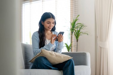 Oturma odasında komik mobil uygulamalar kullanarak rahat bir koltukta oturan gülümseyen genç Asyalı kadın. Evdeki Kadın, Çevrimiçi Alışveriş Yapıyor, Arkadaşlarına Mesajlaşıyor, Sosyal Medya 'ya Gönderiyor