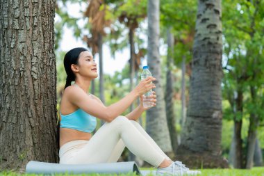 Aktif giysili genç bir Asyalı kadın bir ağacın altında oturmuş parkta spor yaptıktan sonra su içiyor. Rahatlamış ve rahatlamış görünüyor. Açık havada huzurlu bir ortamda susuzluktan ve doğadan hoşlanıyor.