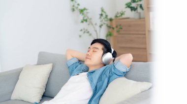 Rahat genç Asyalı adam rahat koltukta dinleniyor, sakin çekici adam kanepede dinleniyor, temiz hava soluyor gözleri kapalı, kulaklık takıyor.