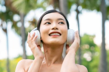 Asyalı sporcu kadın dışarıda kulaklıkla müzik dinliyor. Spor giyimli bir kadın parkta ayakta duruyor. Mutlu bayan sporcu antrenmanına ara veriyor.