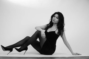 Stüdyoda beyaz arka planda yüksek topuklu giysili, küçük siyah elbiseli seksi esmer kadın. 