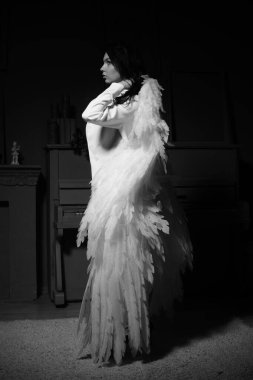 Güzel esmer kadın spor ceketli ve beyaz melek kanatlarıyla stüdyoda piyanonun yanında poz veriyor. 