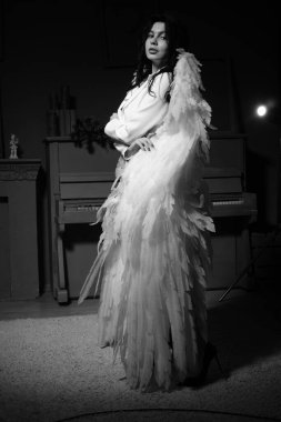 Güzel esmer kadın spor ceketli ve beyaz melek kanatlarıyla stüdyoda piyanonun yanında poz veriyor. 