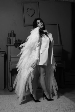 Güzel esmer kadın spor ceketli ve beyaz melek kanatlarıyla stüdyoda piyanonun yanında poz veriyor. 