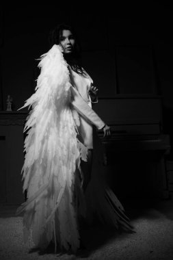 Güzel esmer kadın spor ceketli ve beyaz melek kanatlarıyla stüdyoda piyanonun yanında poz veriyor. 