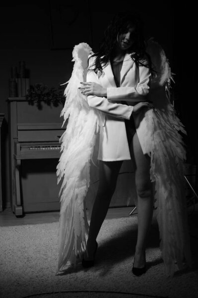 Beautiful brunette woman in blazer and white angel wings posing near piano at studio