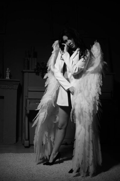Beautiful brunette woman in blazer and white angel wings posing near piano at studio