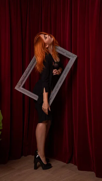 young Red-haired woman posing with empty antique frames on red background