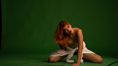 Stüdyoda beyaz gömlek ve külot giyen, yeşil duvar arkasında poz veren çekici, çıplak genç bir kadın.  