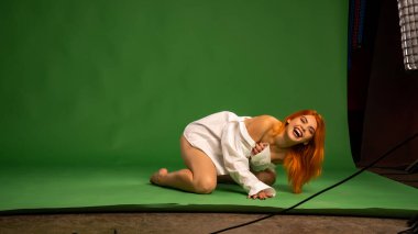 Stüdyoda beyaz gömlek ve külot giyen, yeşil duvar arkasında poz veren çekici, çıplak genç bir kadın.  