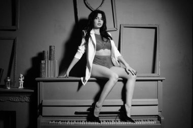 Beyaz ceketli ve iç çamaşırlı güzel esmer kadın stüdyoda piyanoyla poz veriyor. 