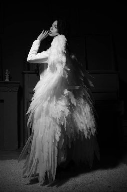 Güzel esmer kadın spor ceketli ve beyaz melek kanatlarıyla stüdyoda piyanonun yanında poz veriyor. 