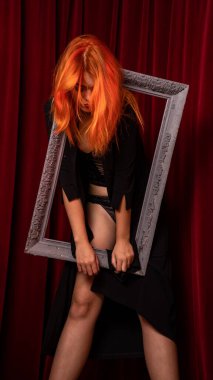 Kızıl saçlı genç kadın kırmızı arka planda boş antika çerçevelerle poz veriyor. 