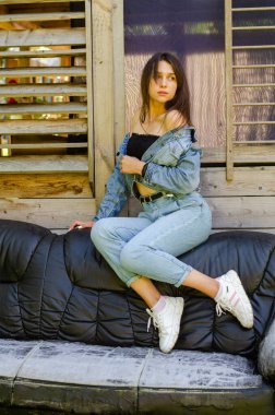 Yazın güneşli bir günde evin yanındaki deri koltukta poz veren kot elbiseli güzel bir genç kız. 