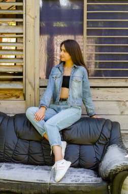 Yazın güneşli bir günde evin yanındaki deri koltukta poz veren kot elbiseli güzel bir genç kız. 