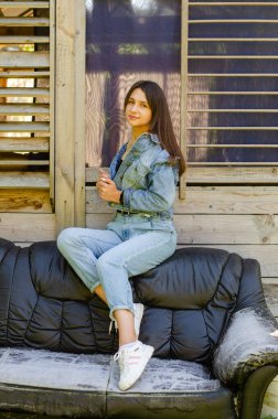 Yazın güneşli bir günde evin yanındaki deri koltukta poz veren kot elbiseli güzel bir genç kız. 