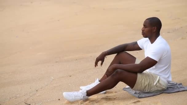 Afro-americano desportivo homem sentado na praia e olha para o mar — Vídeo de Stock