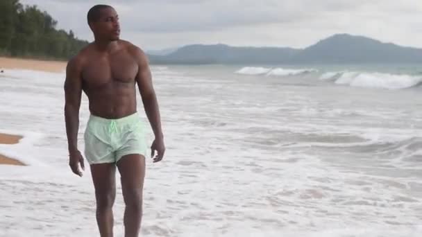 Homem afro-americano caminha na praia ao pôr-do-sol. shorts e top nu — Vídeo de Stock