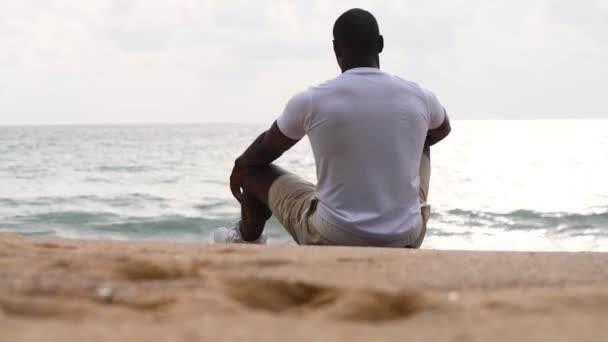 Afro Amerikaanse sportieve man zit op het strand en kijkt naar de zee Rechtenvrije Stockvideo