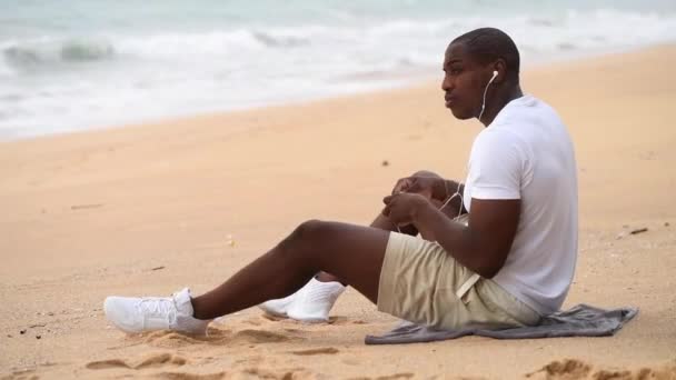 Afro-americano desportivo homem sentado na praia e olha para o mar e ouvir música — Vídeo de Stock