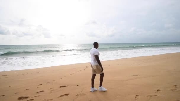 Afro americano desportivo cara caminha ao longo da praia e olha para o mar e ouvir música — Vídeo de Stock