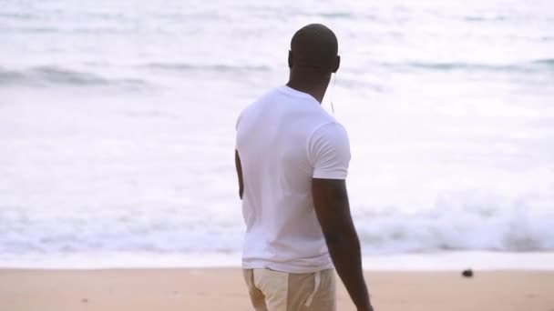 Afro americano desportivo cara caminha ao longo da praia e olha para o mar e ouvir música — Vídeo de Stock