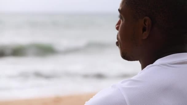 Afroamerikanischer sportlicher Mann sitzt am Strand und blickt aufs Meer — Stockvideo