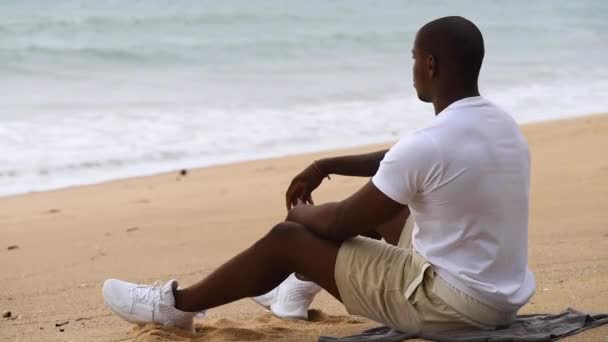 Afro-americano desportivo homem sentado na praia e olha para o mar — Vídeo de Stock