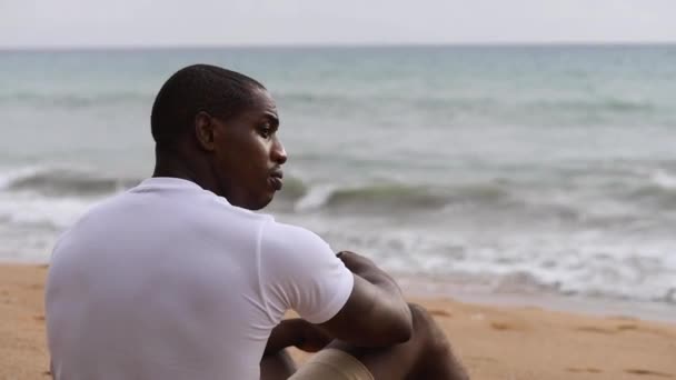 Afro Amerikaanse sportieve man zit op het strand en kijkt naar de zee Rechtenvrije Stockvideo