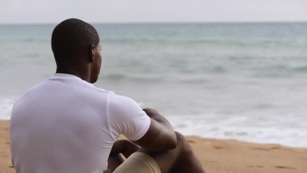 ビーチに座って海を見ているアフリカ系アメリカ人のスポーティな男 — ストック動画