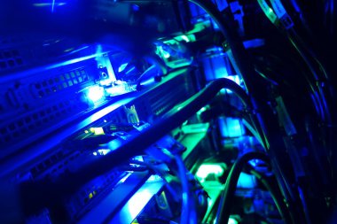 Bu resim aydınlanmış bilgisayar sunucularını canlı mavi ve yeşil ışıklarla yakalar, modern bir veri merkezinde karmaşık kablolar ve elektronik bileşenler gösterir.