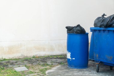 Mavi bir çöp kutusu ve siyah çöp torbalarıyla dolu daha büyük bir çöp konteynırı bir duvara yaslanmış, kentsel atık yönetimi ve çevre bakımını gözler önüne seriyor..