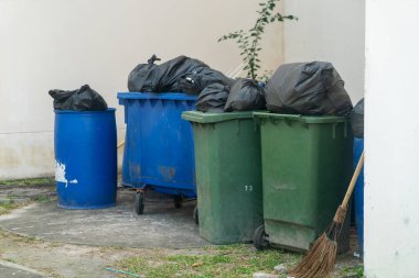 Kentsel bir açık alanda çöp bidonları ve siyah çöp torbaları koleksiyonu, atık yönetimi ve çevrede temizlik ihtiyacı gösteriliyor.