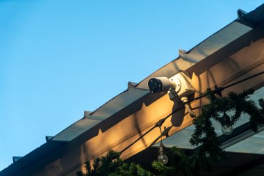 Çatıya monte edilmiş bir güvenlik kamerası açık mavi bir gökyüzü yakalıyor ve kentsel ortamlarda güvenlik ve izleme için kullanılan modern izleme teknolojisini gösteriyor..